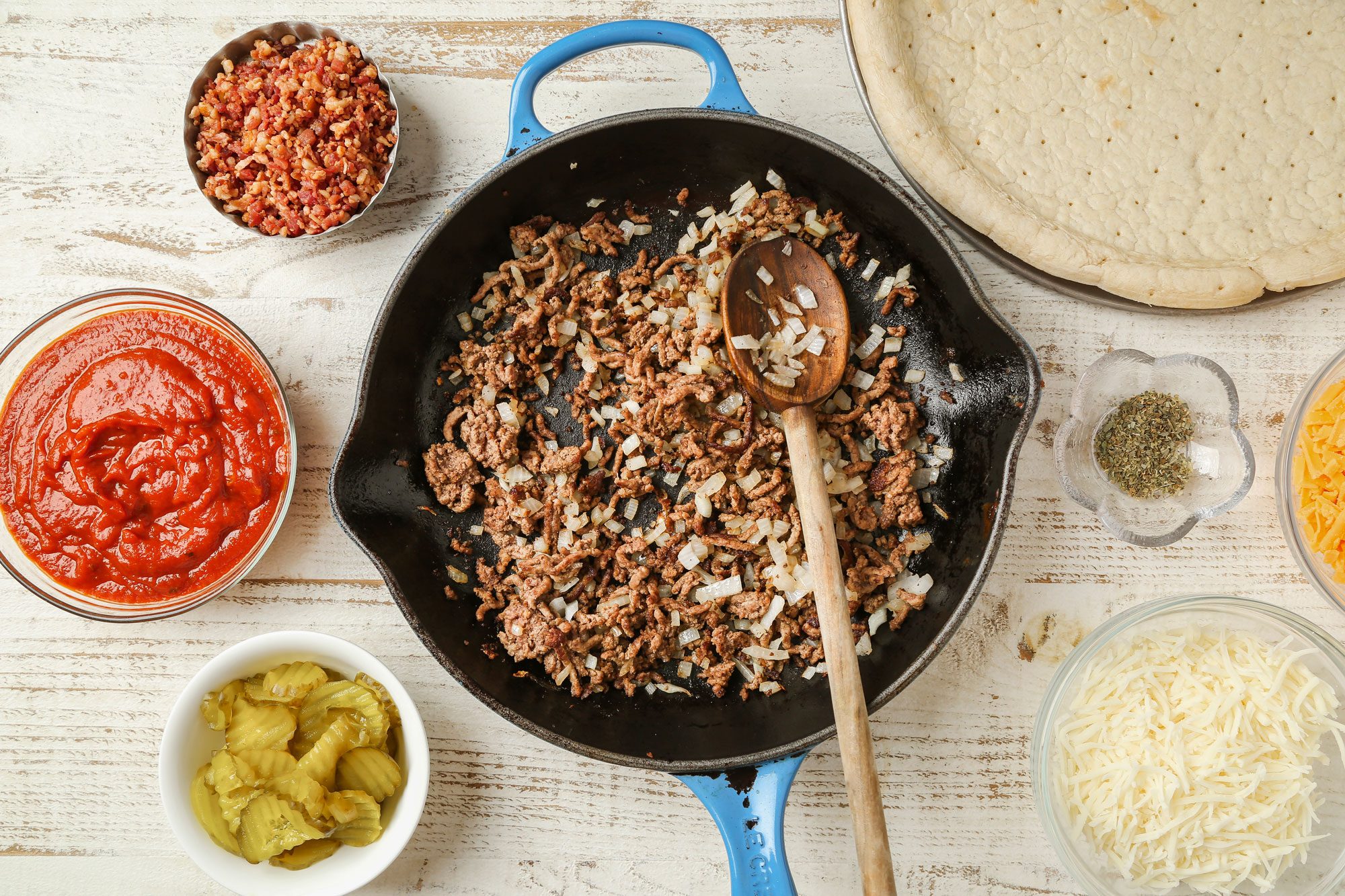Top Shot of a skillet of cooked ground beef and onions is surrounded by pizza sauce; chopped bacon; shredded cheese; pickles; dried herbs; and an uncooked pizza crust on a wooden table background