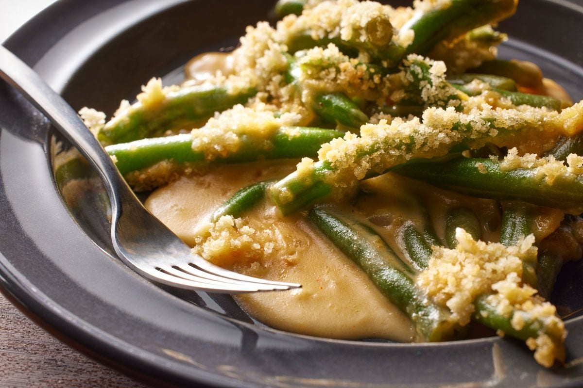A closeup shot of Beer Cheese Green Bean Casserole with creamy sauce and crispy breadcrumbs, on a dark plate with a fork