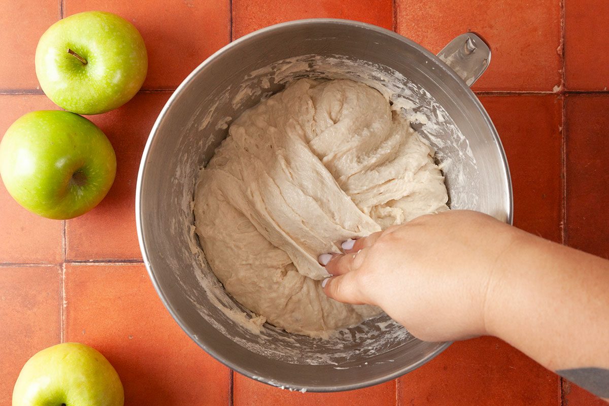 Overhead shot of a hand stretching dough inside a metal mixing bowl on a tiled surface, with two green apples nearby;