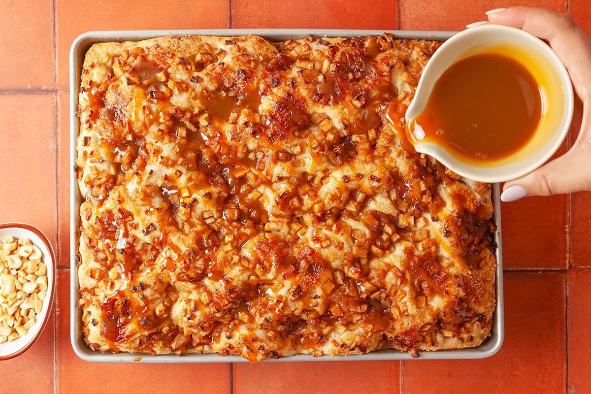 Overhead shot of a hand pouring caramel sauce over a freshly baked sheet cake topped with chopped nuts, on a tiled surface; with a small bowl of extra nuts to the side.