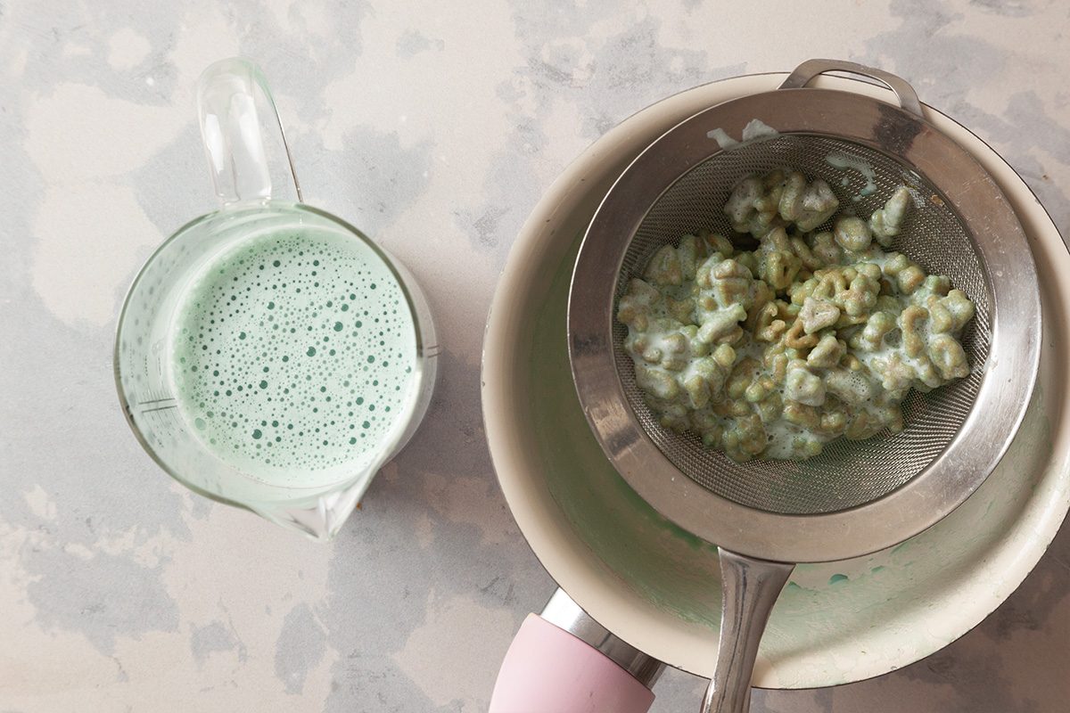A metal strainer holding green clumps rests over a white bowl, while a measuring cup nearby contains a frothy green liquid on a light-colored surface.