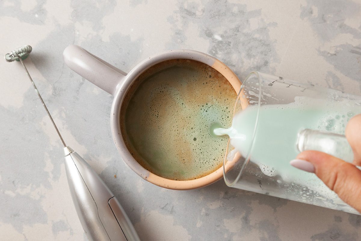 A hand pours frothed milk from a small pitcher into a mug of coffee on a light-colored surface, with a handheld milk frother next to the mug.