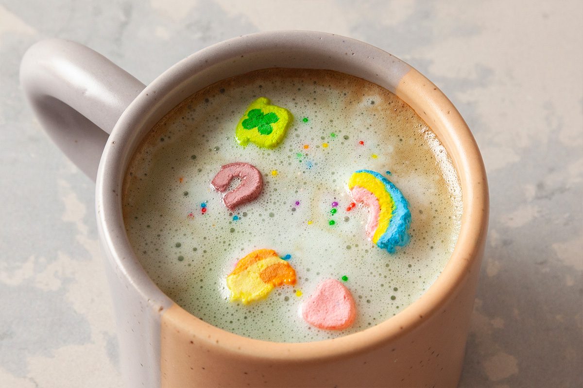 A mug of frothy milk with colorful marshmallow shapes floating on top, resembling clovers, hearts, moons, and rainbows.