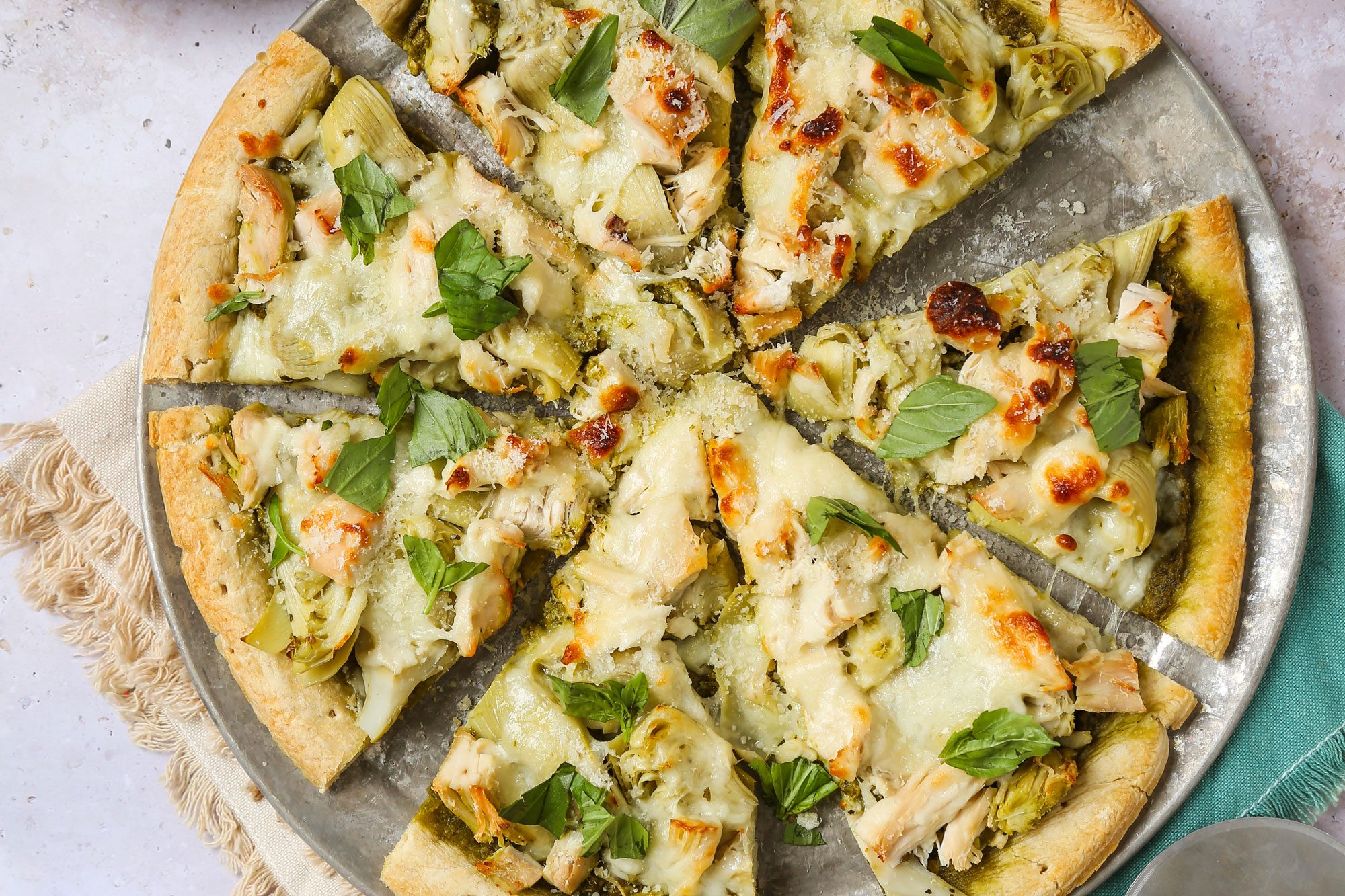 overhead vertical shot of a sliced artichoke pizza topped with fresh basil on a round pan, with a pizza cutter, a plate with pizza slice, a bowl of grated cheese, fresh basil leaves, and a bowl of artichokes nearby on a light surface