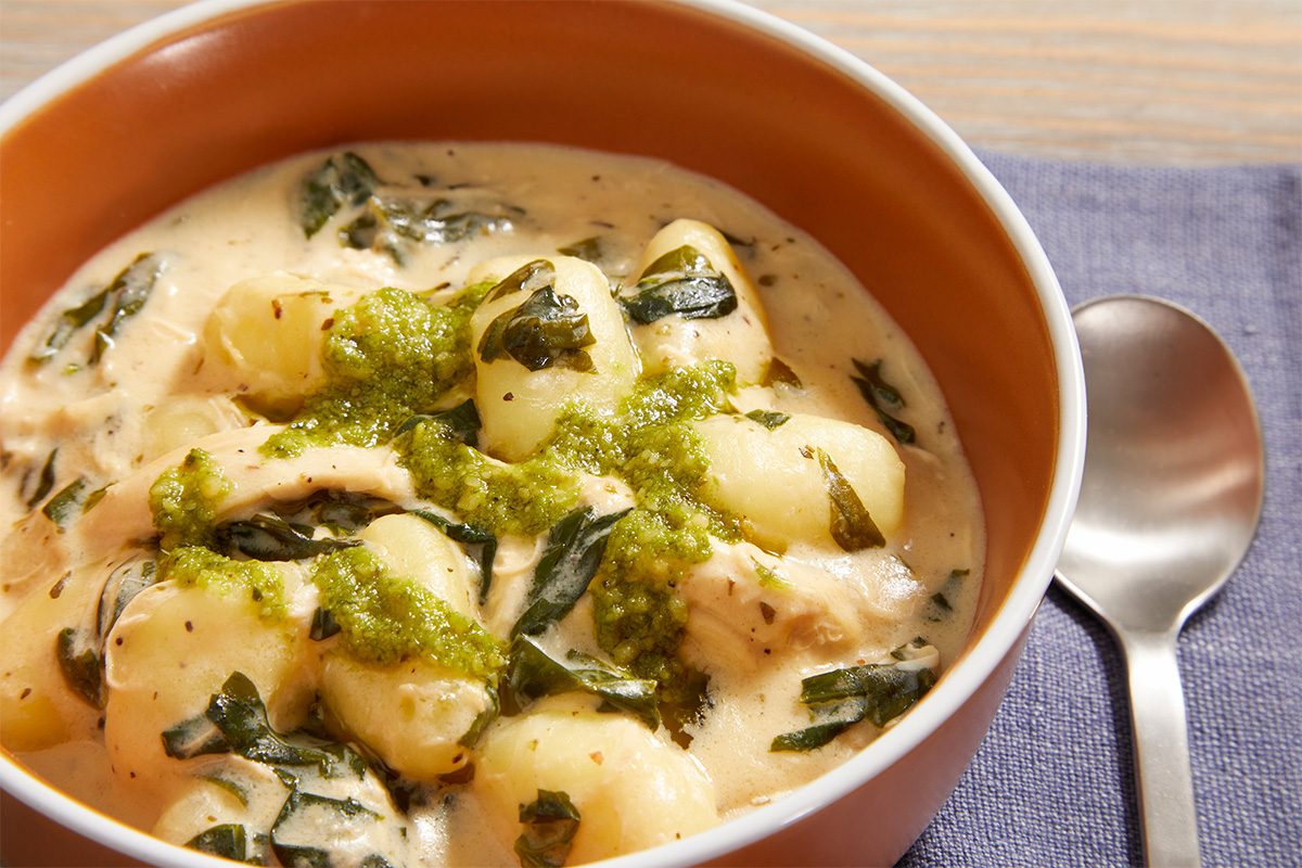 A bowl of creamy gnocchi soup with spinach and shredded chicken, topped with a swirl of green pesto, sits on a purple napkin beside a silver spoon.
