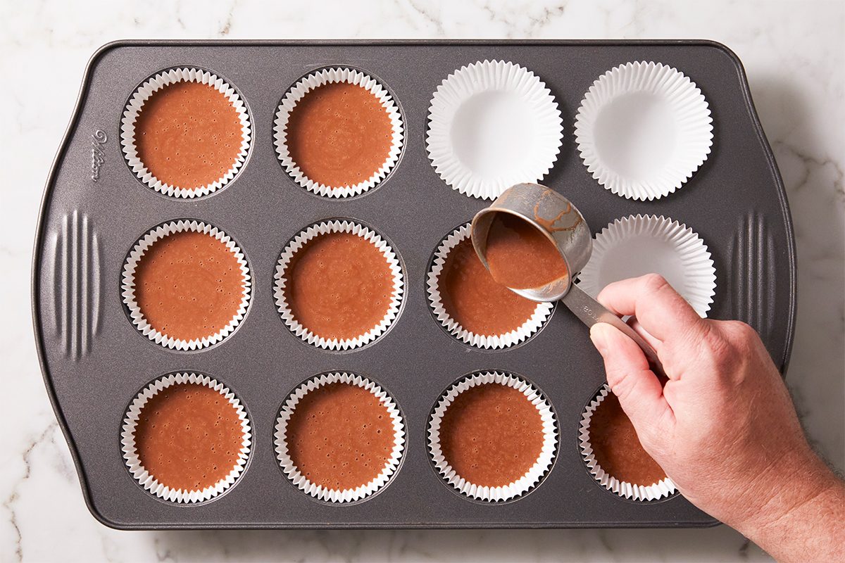 A hand pours chocolate cupcake batter from a measuring cup into paper liners arranged in a muffin tin on a marble countertop.