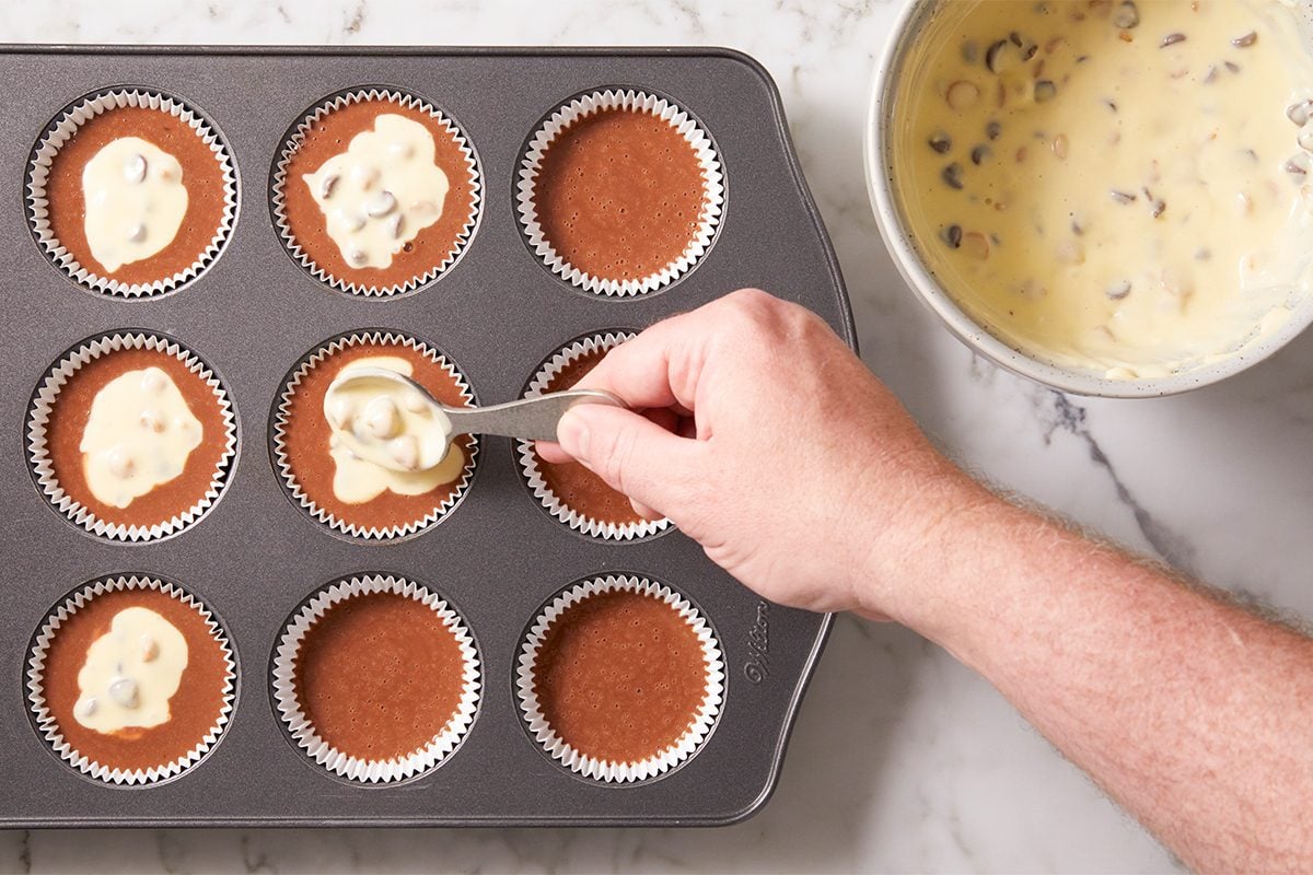 A hand uses a spoon to add a light-colored batter with chocolate chips into cupcake liners filled with a darker batter in a muffin tray. A bowl of chip-filled batter sits nearby on a marble countertop.