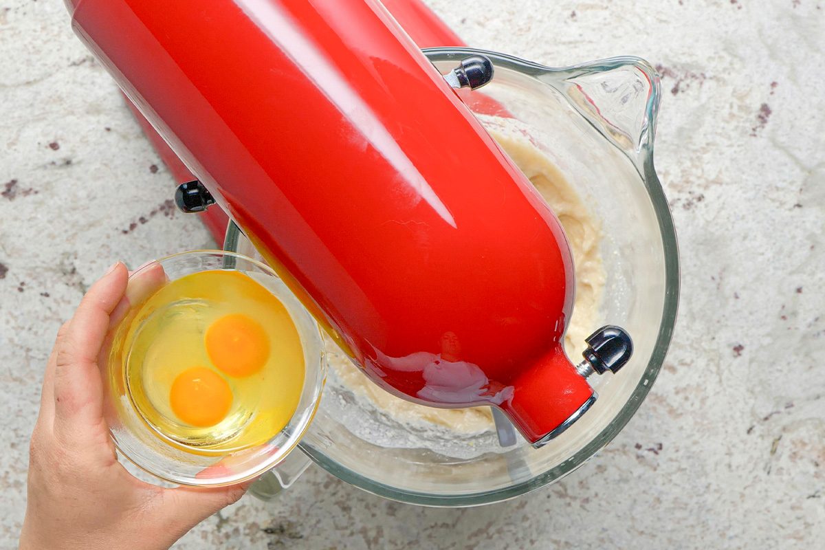 A hand holds a small bowl with two cracked eggs above a red stand mixer containing batter, ready to add the eggs. The scene is set on a light-colored countertop.