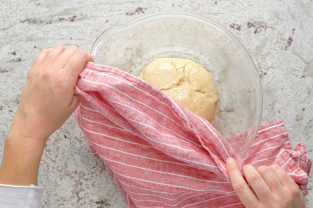 A person covers a glass bowl of dough with a red and white striped cloth on a light countertop.