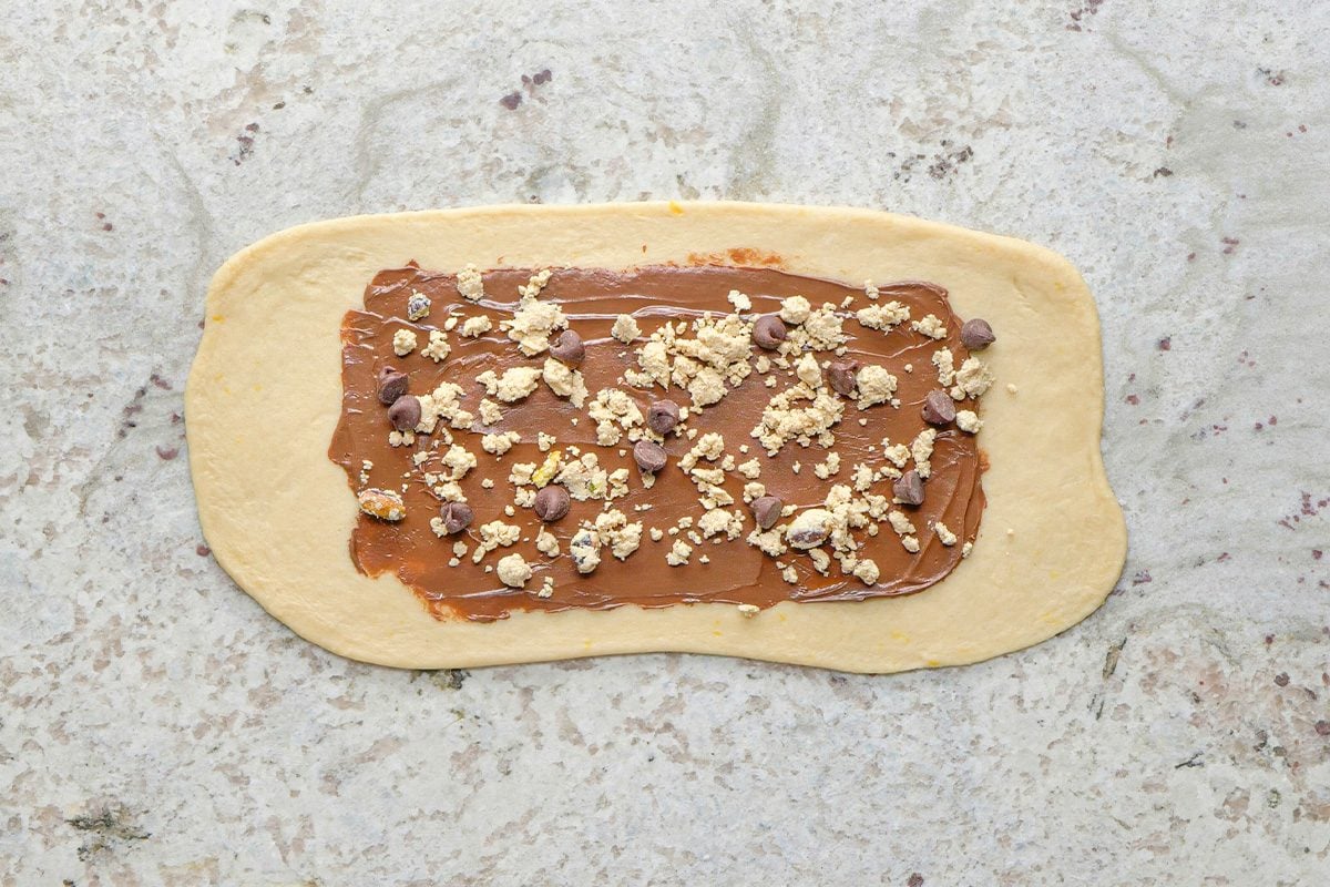 A rectangle of dough on a light countertop is spread with chocolate hazelnut spread and sprinkled with chopped nuts and chocolate chips.