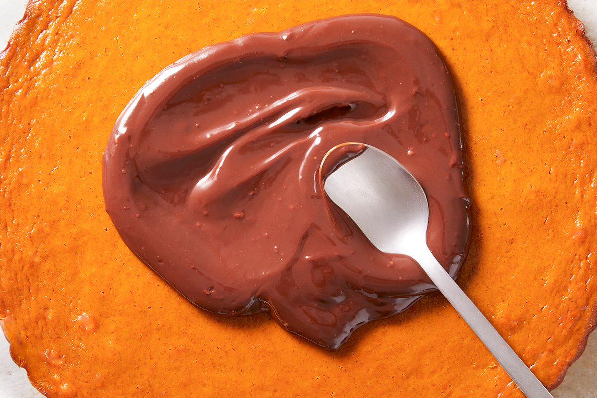 A silver spoon spreads smooth melted chocolate over a round, golden-brown baked crust.