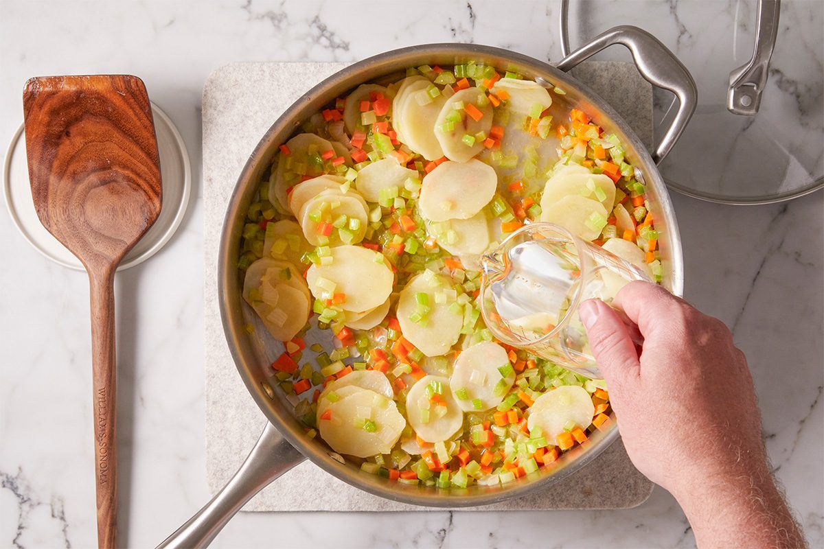 A hand pours liquid into a pan filled with sliced potatoes, diced carrots, celery, and onions on a marble countertop. A wooden spatula rests nearby on a coaster.