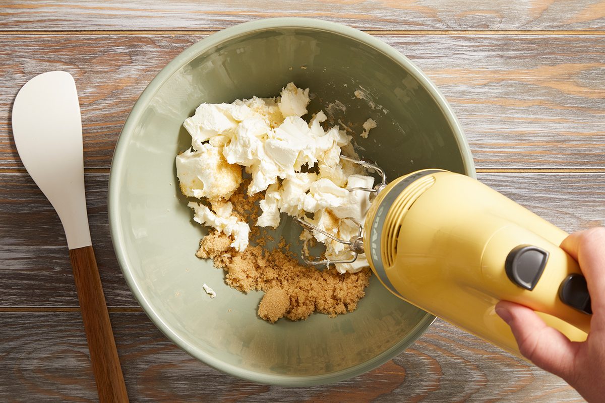 A hand holds a yellow electric mixer above a green bowl containing cream cheese and brown sugar, with a white spatula resting nearby on a wooden surface.