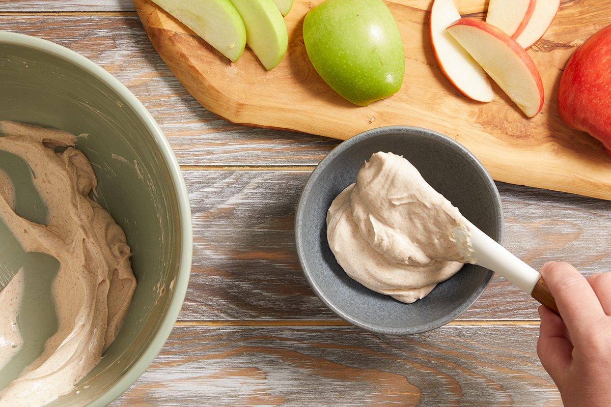 A person scoops creamy dip into a small bowl with a spatula. Nearby, sliced green and red apples are arranged on a wooden board, and a larger mixing bowl sits to the side. All items rest on a wooden surface.