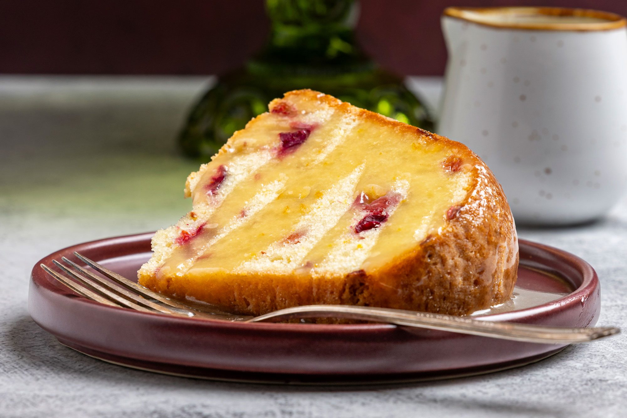 Table view of a Cranberry-Orange Pound Cake displayed on a green glass cake stand; A single slice is served on a brown plate with a fork; set on a light surface against a deep red background