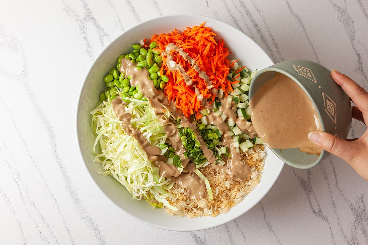Overhead shot of a hand pouring creamy dressing over a bowl filled with shredded cabbage, carrots, edamame, diced cucumber, green onions, and shredded chicken, set on a white marble surface;