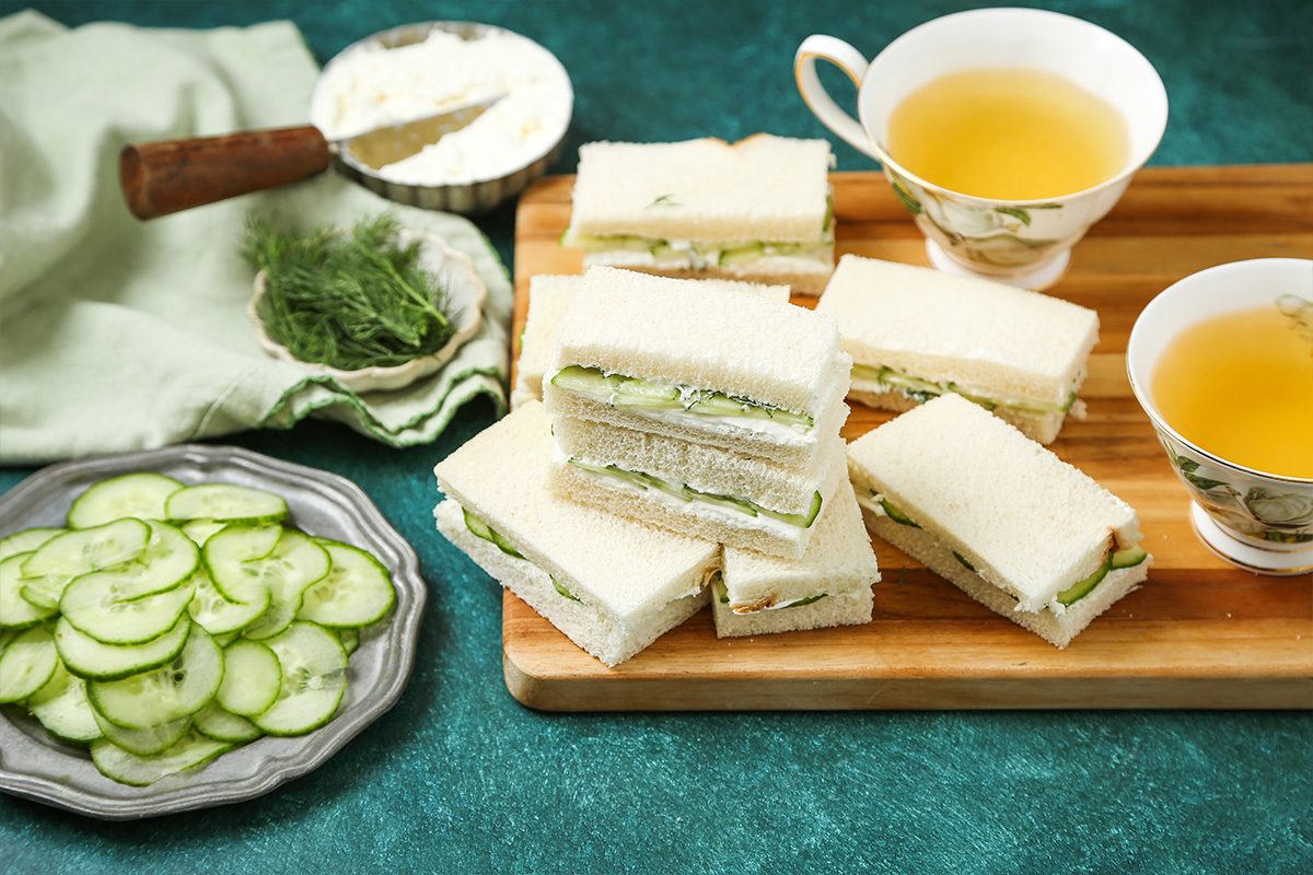 A wooden tray with stacked cucumber tea sandwiches sits next to two cups of tea. Nearby are a plate of sliced cucumbers, a bowl of spread, and fresh dill on a green tablecloth.
