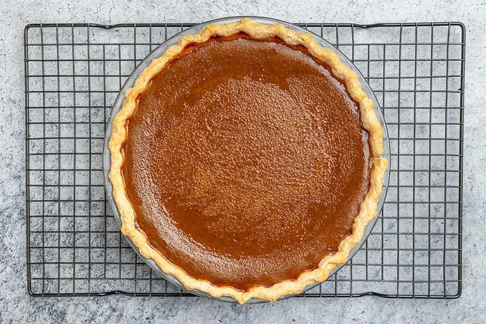 A whole pumpkin pie with a golden, flaky crust sits on a round metal cooling rack, viewed from above against a gray textured background.