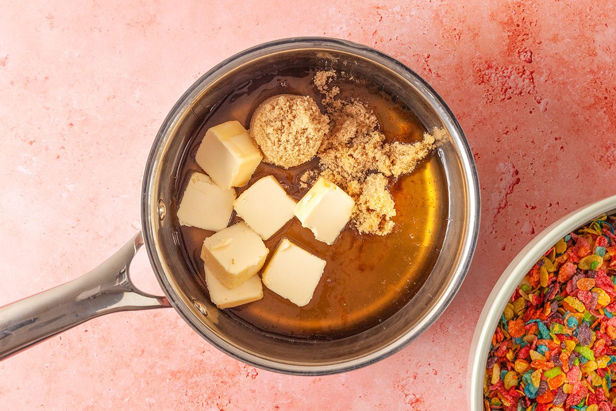 Overhead shot of a saucepan with cubed butter and brown sugar on a pink surface, next to a bowl of colorful cereal pieces
