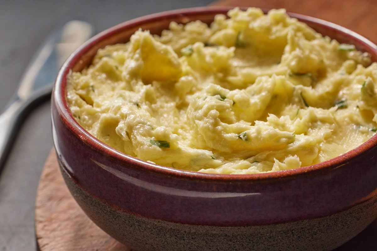 3/4th closeup shot of A rustic ceramic bowl of Garlic Chive Butter mashed potatoes garnished with chopped chives, sitting on a wooden surface
