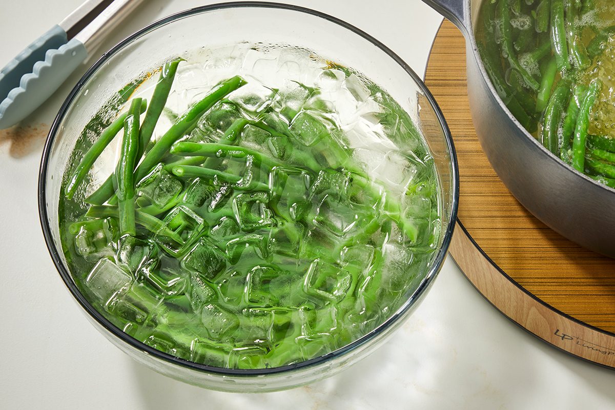 A glass bowl filled with green beans and ice cubes sits on a white surface next to a pot of cooked green beans on a wooden trivet. Tongs are placed nearby.