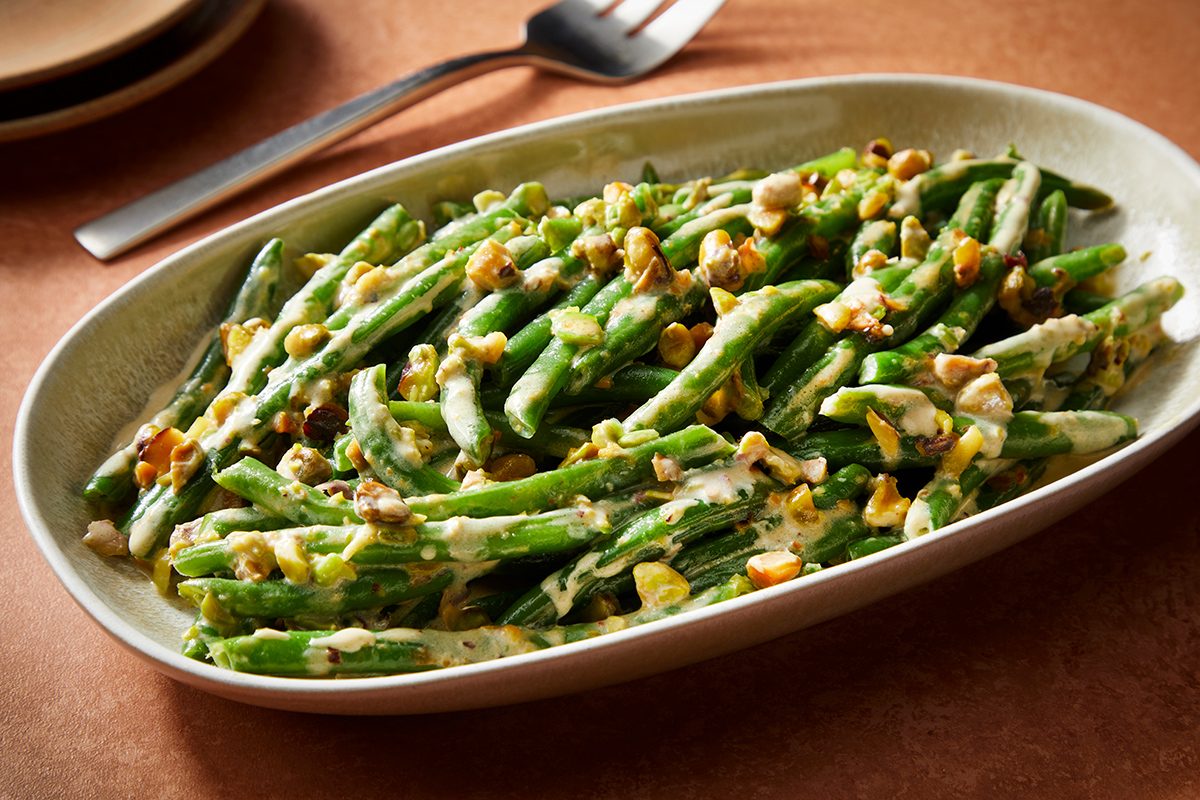 A serving dish filled with green beans topped with creamy sauce and sprinkled with chopped nuts, set on a brown table with a fork and plates in the background.