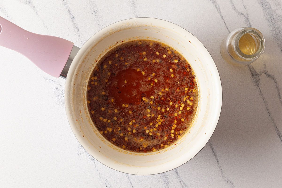 A saucepan with a pink handle contains a bubbling mixture of sauce with visible red chili flakes and seeds. Beside it, there is a small glass bottle on a white marble countertop.