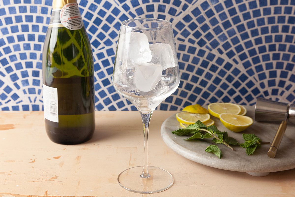 A wine glass with ice cubes sits on a table next to a bottle of sparkling wine, lemon slices, fresh mint, and a cocktail shaker, with a blue and white tiled background.