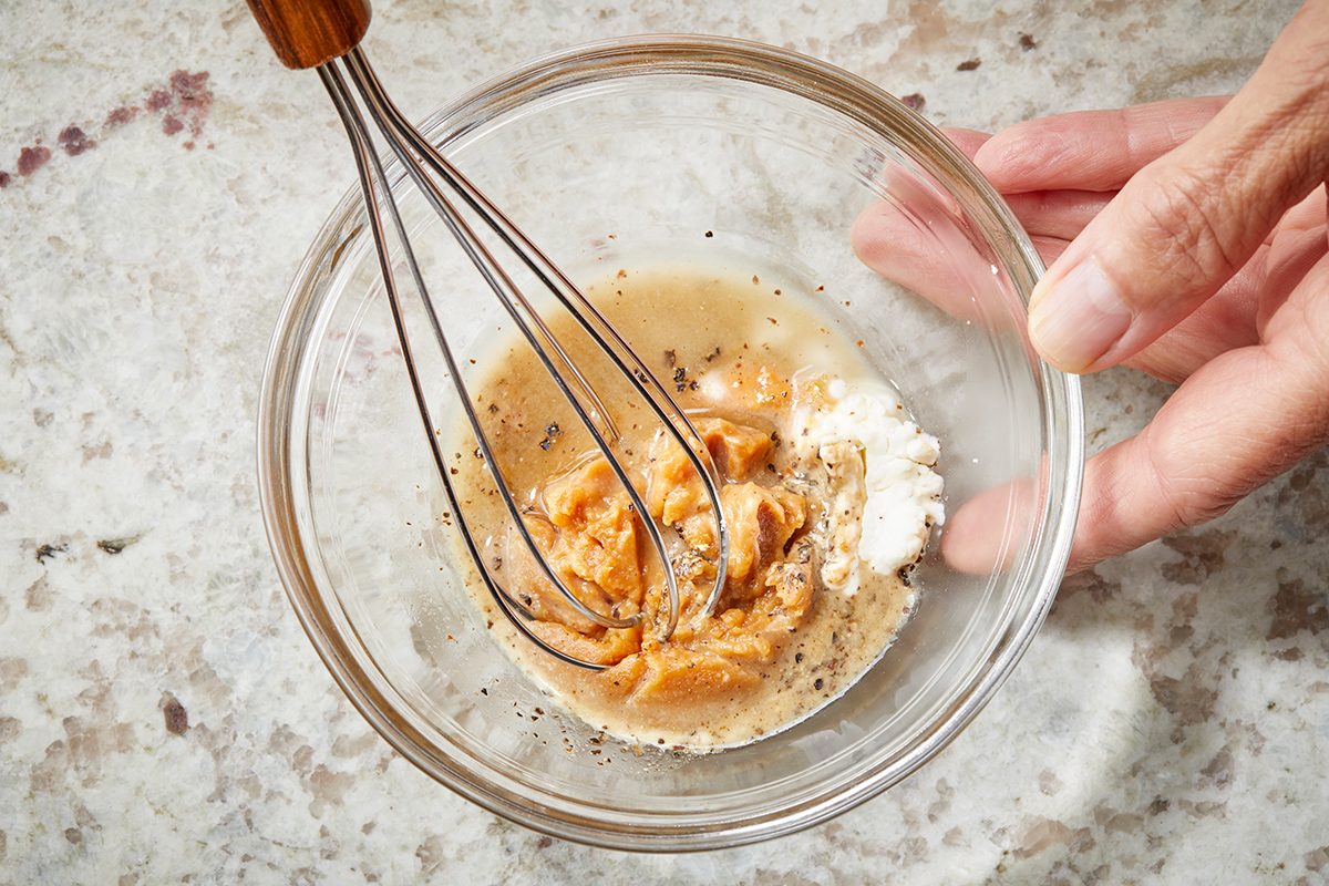 A hand holds a glass bowl with a whisk mixing peanut butter, flour, and liquid ingredients on a light speckled countertop.