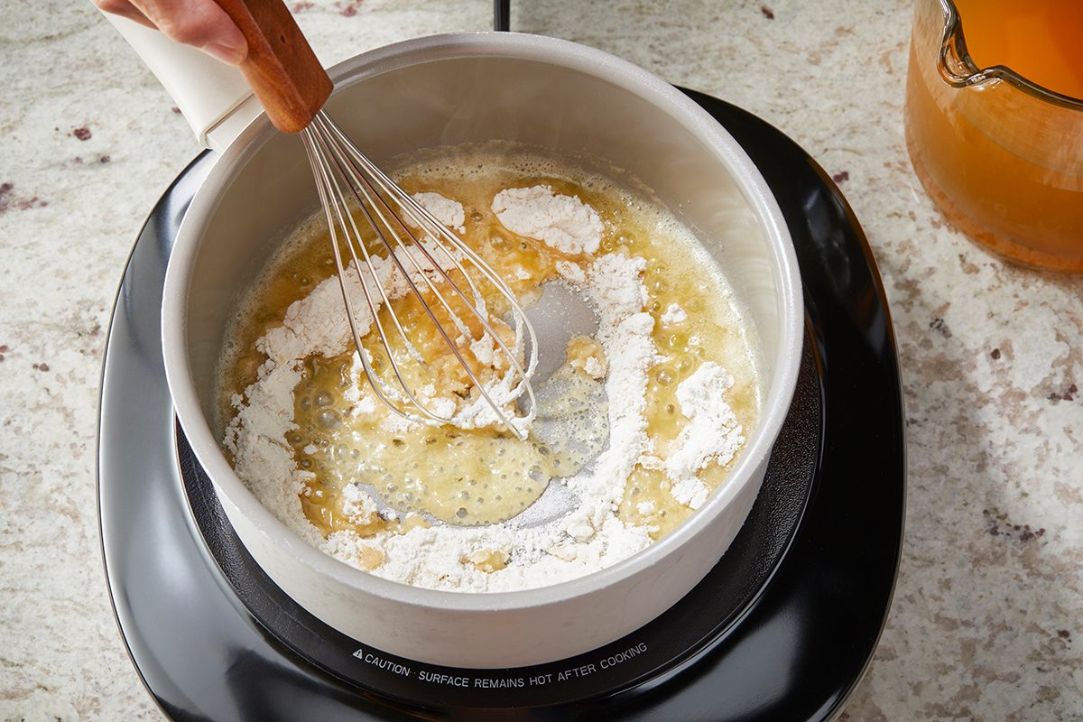 A person uses a whisk to mix flour and melted butter in a saucepan on a stovetop, preparing a roux. A jug with a golden liquid is visible nearby on a marbled countertop.