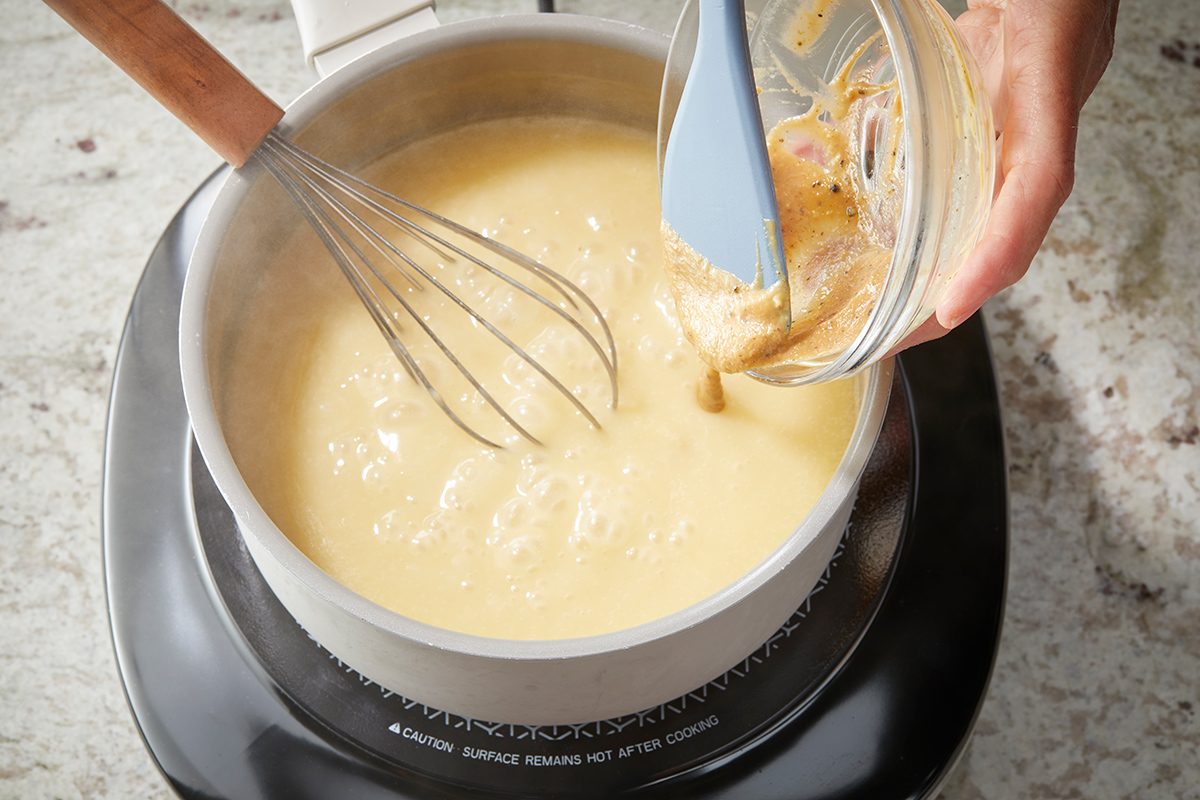 A hand pours a mixture from a small bowl into a pot of thick, creamy sauce on a stovetop. A whisk rests in the pot, and the scene is set on a marbled countertop.