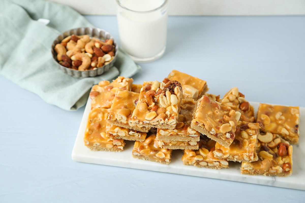 A stack of nut brittle bars with visible peanuts and cashews sits on a white tray, next to a small bowl of mixed nuts, a folded blue napkin, and a glass of milk on a light blue surface.