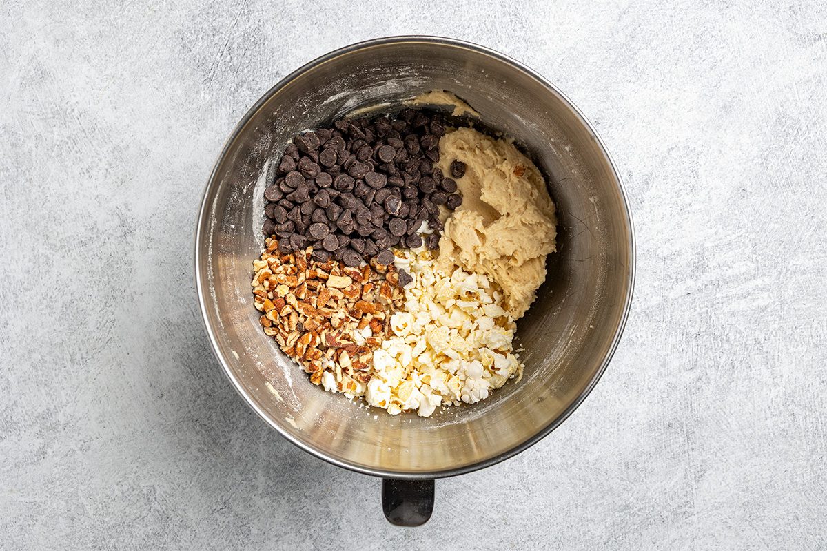 A metal mixing bowl containing cookie dough, chocolate chips, chopped pecans, and popcorn, each ingredient grouped separately and unmixed.
