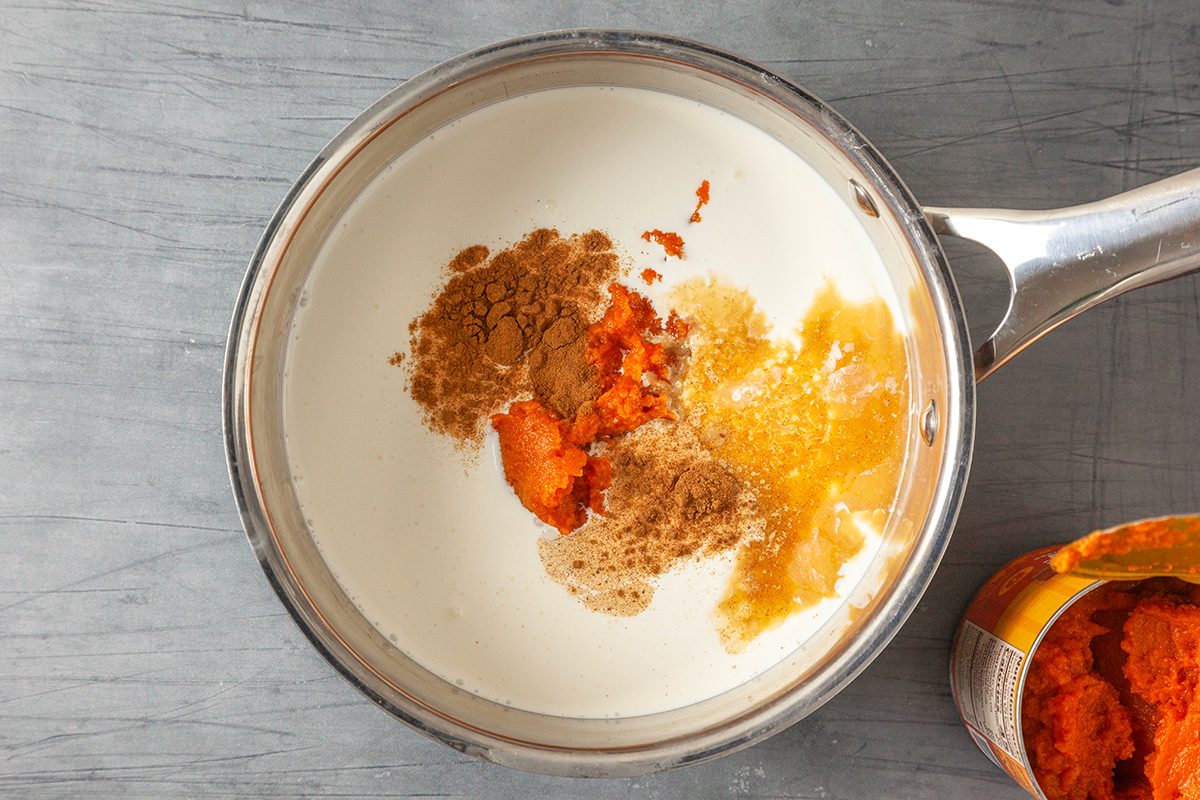 Overhead shot of a saucepan filled with cream, pumpkin purée, brown sugar, and cinnamon, set on a gray surface with an open can of pumpkin purée partially visible beside it