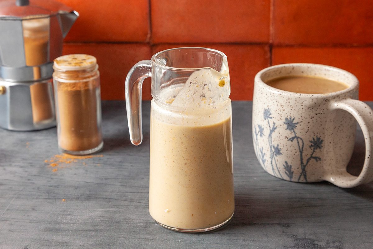 Close of a glass pitcher filled with pumpkin spice creamer, set on a gray surface with a jar of cinnamon and a ceramic mug of coffee in the background against red tiles