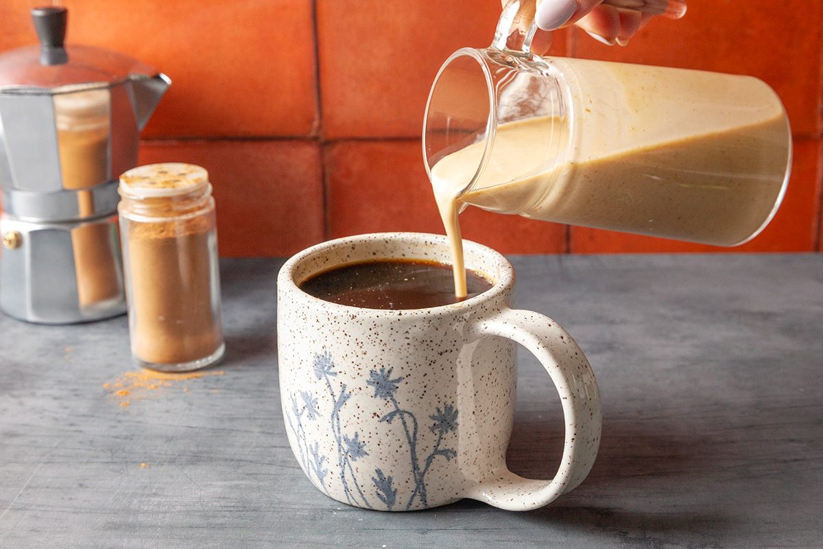 Close-up shot of pumpkin spice creamer being poured from a glass pitcher into a speckled ceramic mug filled with black coffee; set on a gray surface with a jar of cinnamon and a stovetop espresso maker in the background