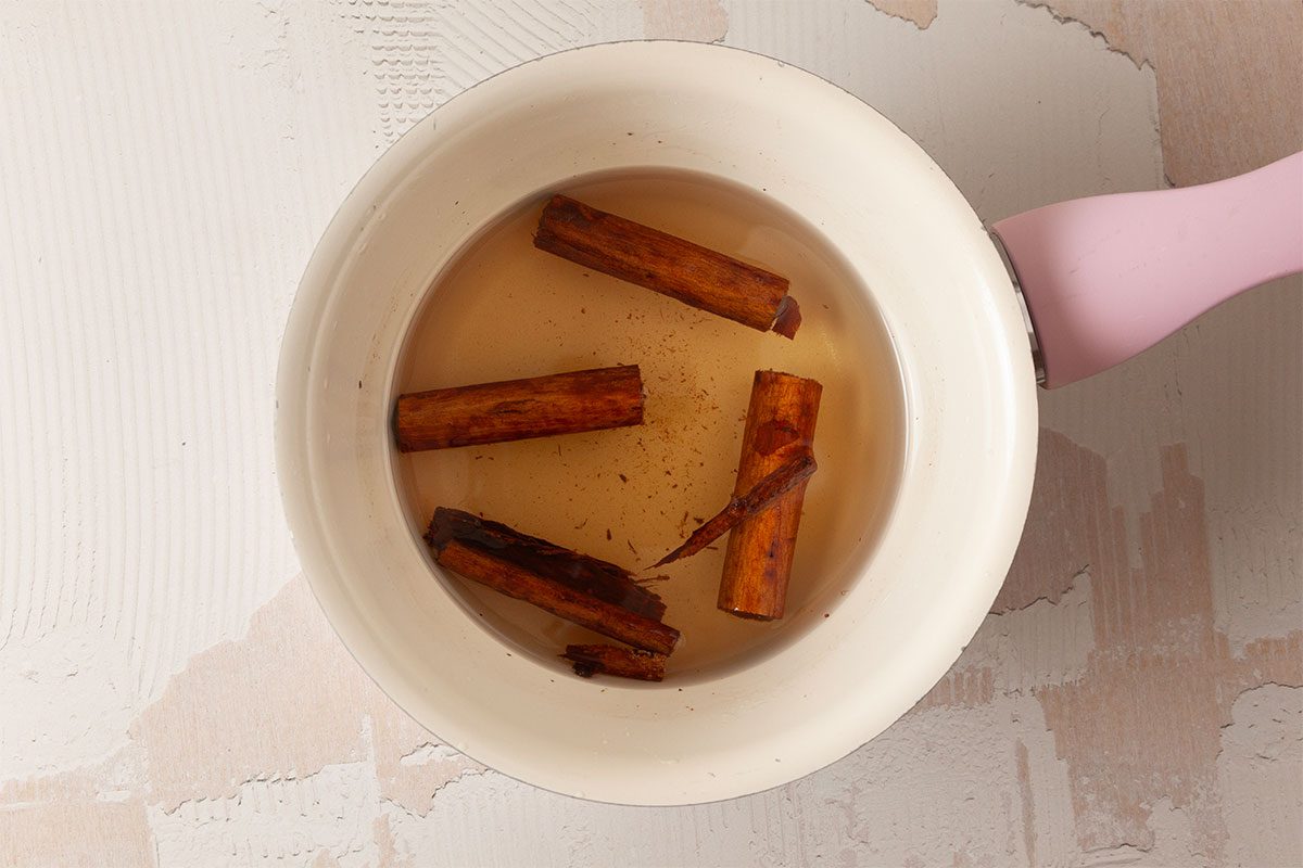A white pot with a pink handle containing water and several cinnamon sticks, placed on a textured, light-colored surface.