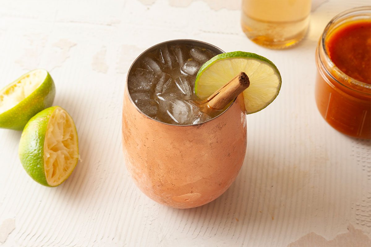 A copper mug filled with ice, garnished with a lime wheel and a cinnamon stick, sits on a light surface. Nearby are squeezed lime halves, a glass of amber liquid, and a jar of red sauce.