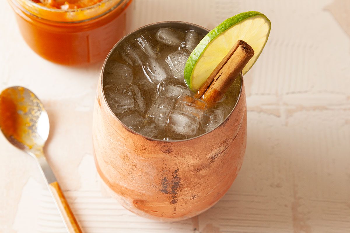 A copper mug filled with ice cubes, garnished with a lime wheel and a cinnamon stick, sits on a light surface near a spoon and a jar containing an orange-colored spread.