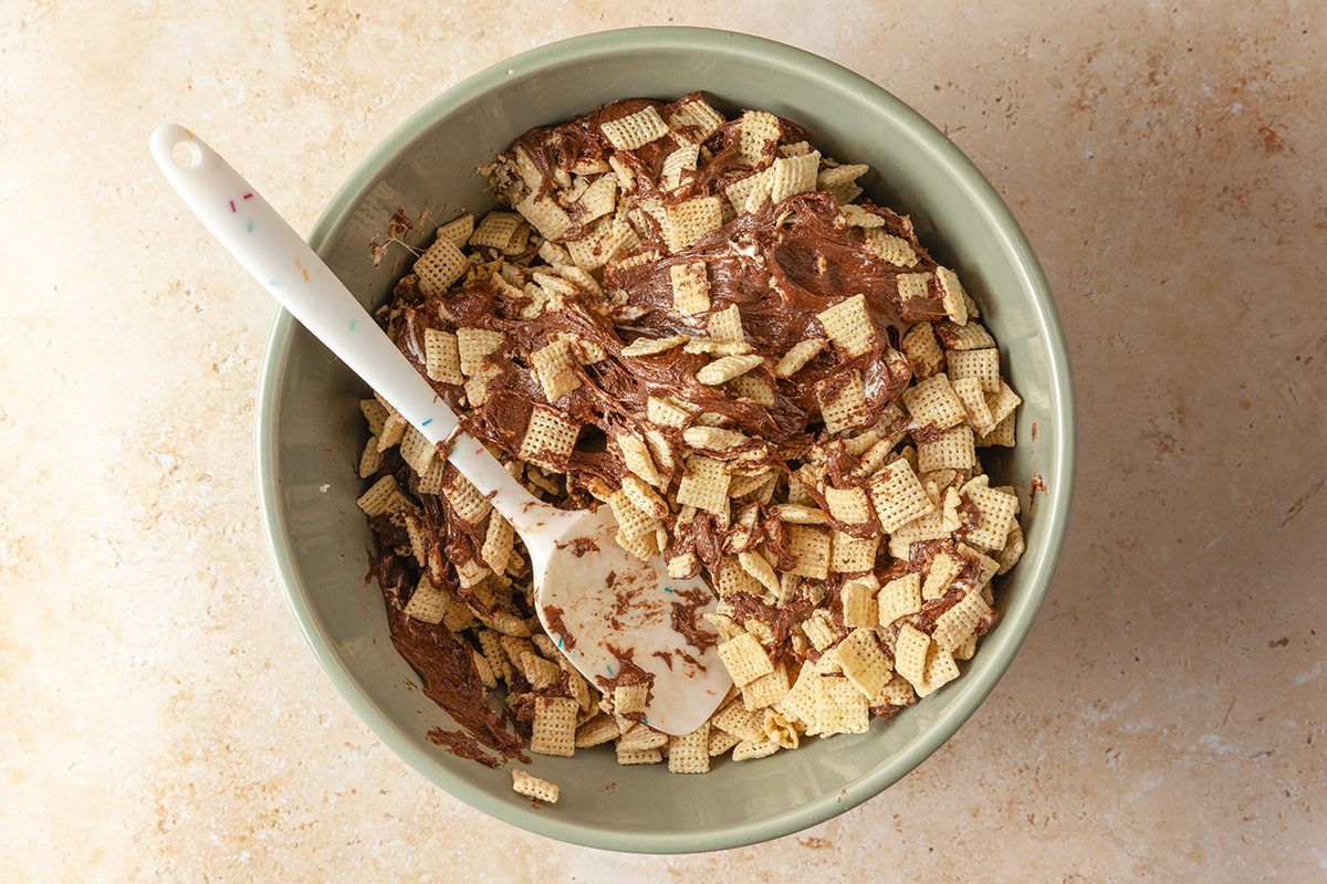 Overhead shot of a green bowl filled with Chex cereal coated in chocolate, with a white spatula resting inside; placed on a light beige countertop