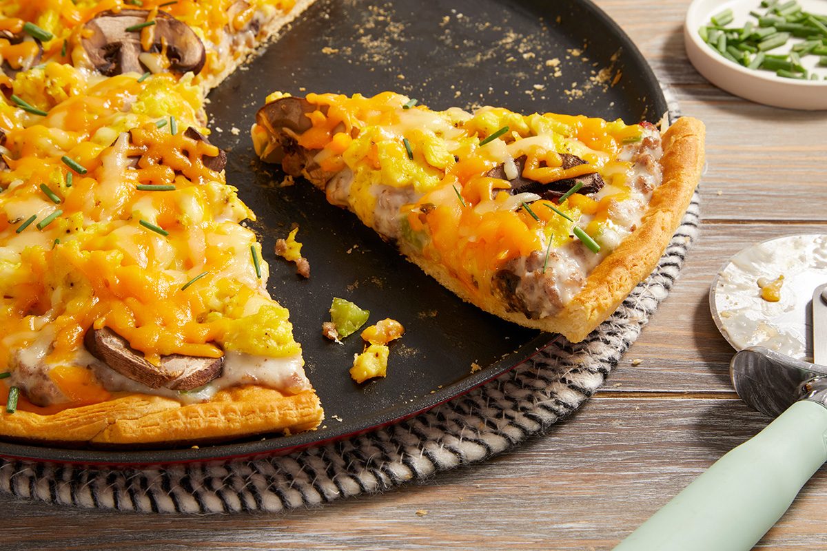 A close-up of a pizza on a black pan with one slice being taken out. The pizza is topped with melted cheese, mushrooms, green peppers, and chives, and sits on a woven mat on a wooden table.
