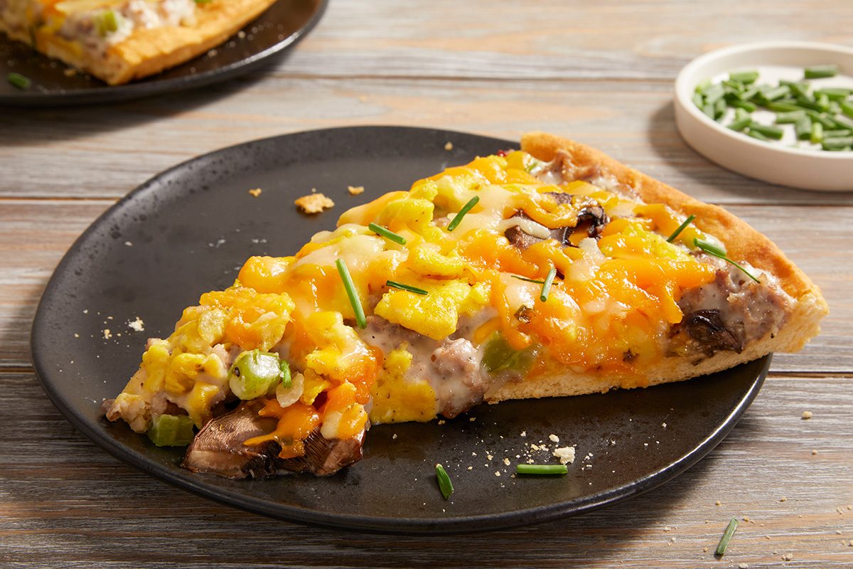 A slice of pizza topped with scrambled eggs, cheese, vegetables, and chives sits on a black plate, with a small dish of chopped chives in the background.