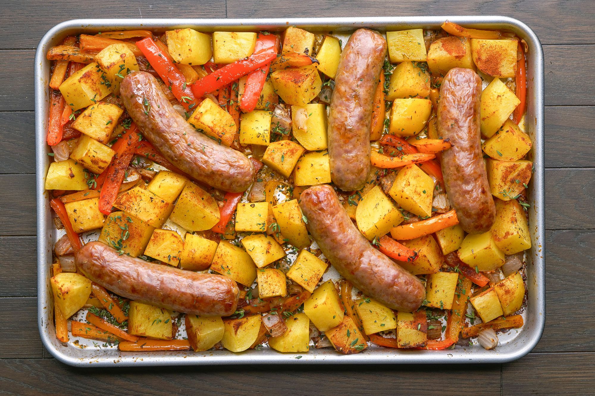 overhead shot of a sheet pan filled with roasted sausages, diced potatoes, and sliced red bell peppers, seasoned with herbs, on a dark wooden surface