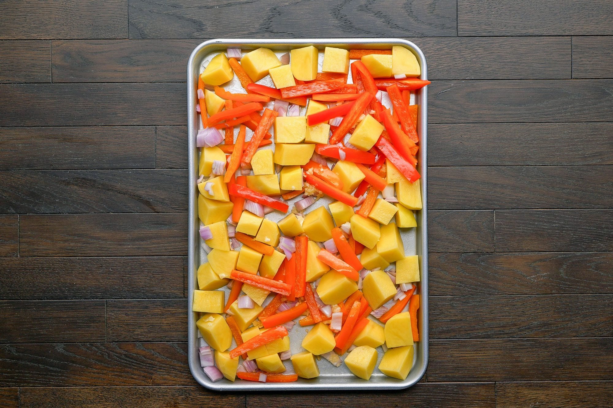 overhead shot of a baking sheet filled with chopped yellow potatoes, sliced red bell peppers, and red onion pieces, set on a dark wooden surface