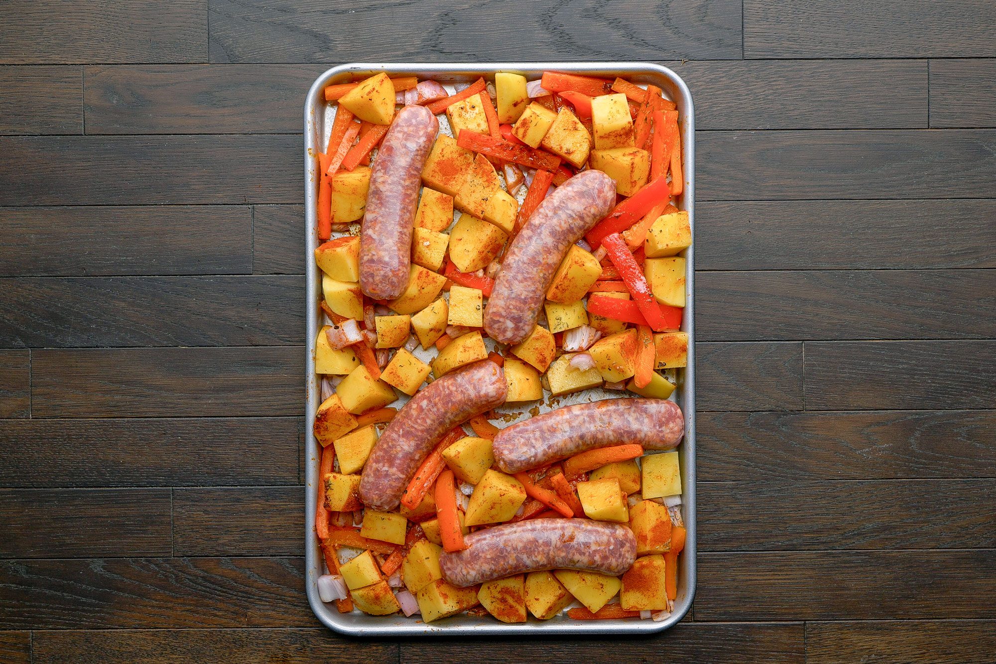 overhead shot of a sheet pan with uncooked sausages, diced potatoes, sliced carrots, and onions, all seasoned and arranged on a dark wooden surface