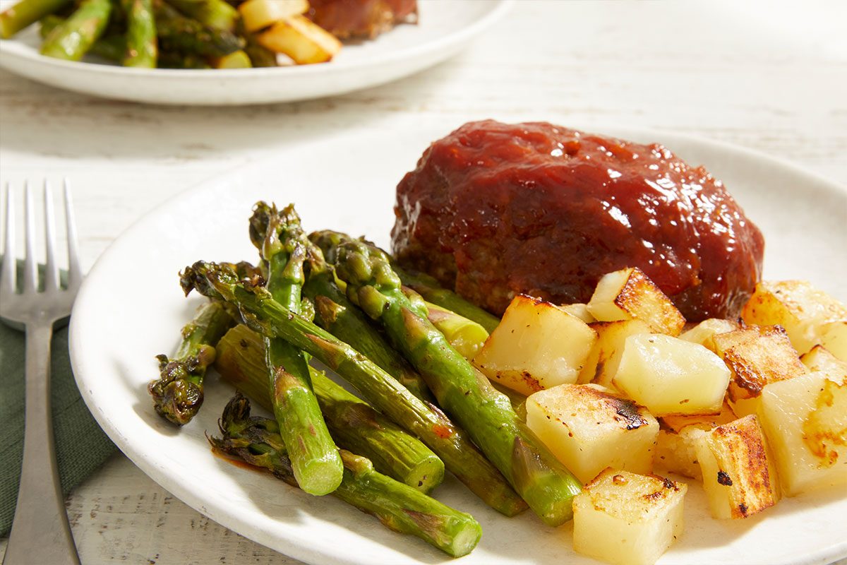 A white plate with a serving of glazed meatloaf, roasted asparagus spears, and golden-brown roasted potatoes, with a fork and another plate in the background.