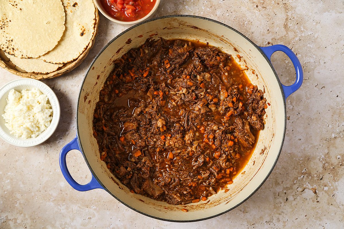 A blue-handled pot filled with shredded beef stew sits on a counter. Nearby are small bowls of shredded cheese, salsa, and a stack of tortillas.