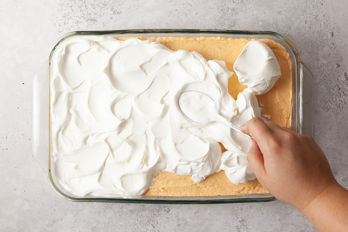 A hand spreading a thick layer of whipped cream over a rectangular yellow cake in a glass baking dish on a light-colored surface.