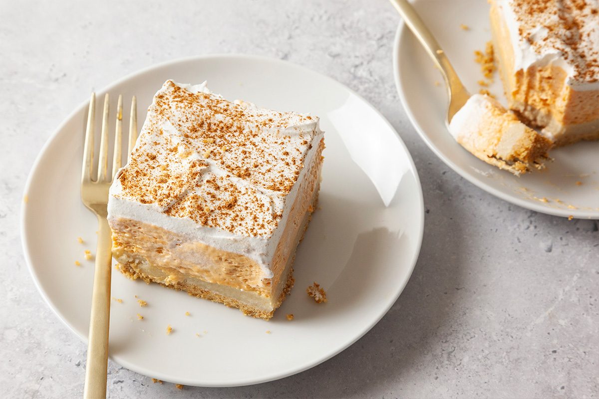 A square slice of creamy dessert with a graham cracker crust and whipped topping sits on a white plate with a gold fork, next to another plate with a partly eaten piece.