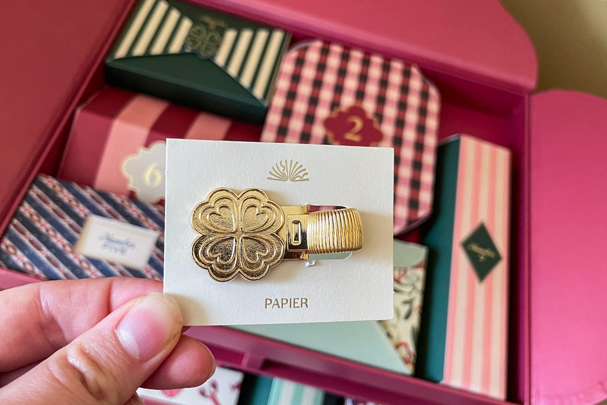 A hand holds a card with two gold paper clips, one shaped like a four-leaf clover and one a rectangular design. In the background, colorful gift boxes with various patterns and numbers are visible.